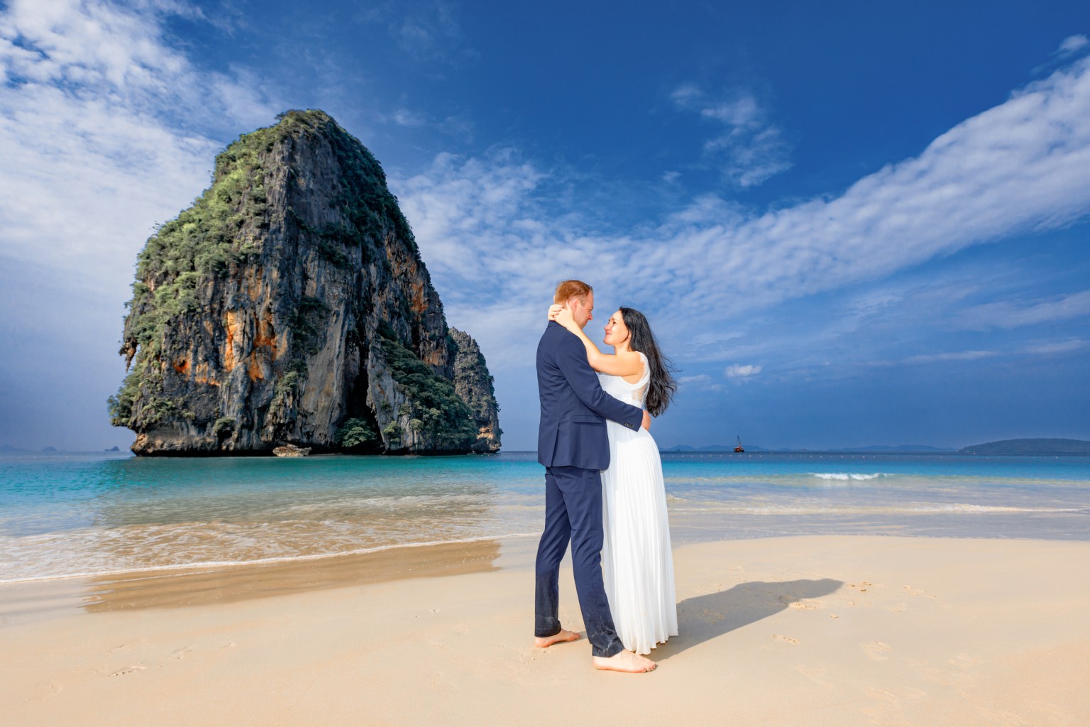 A couple in wedding attire embracing on a sandy beach with a backdrop of a towering limestone cliff and clear turquoise waters under a blue sky.