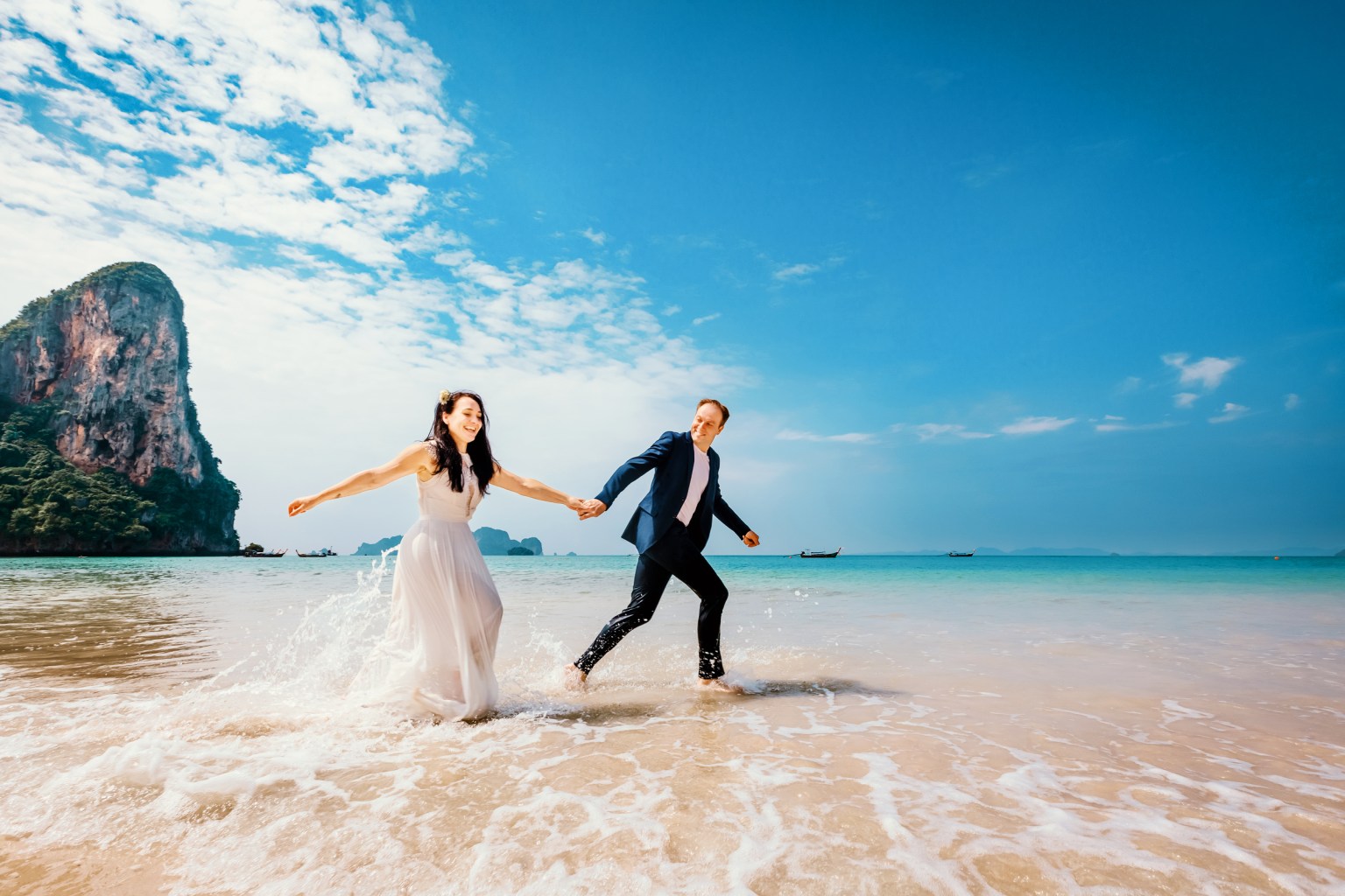 A couple running hand in hand on a sandy beach in Krabi, Thailand, with a limestone cliff in the background and clear blue skies.