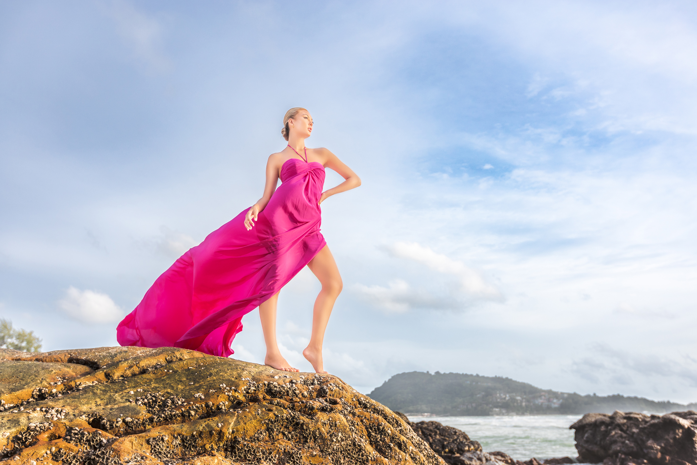 Model wearing a pink dress posing on a rock by the beach with a scenic view of the sky and ocean.