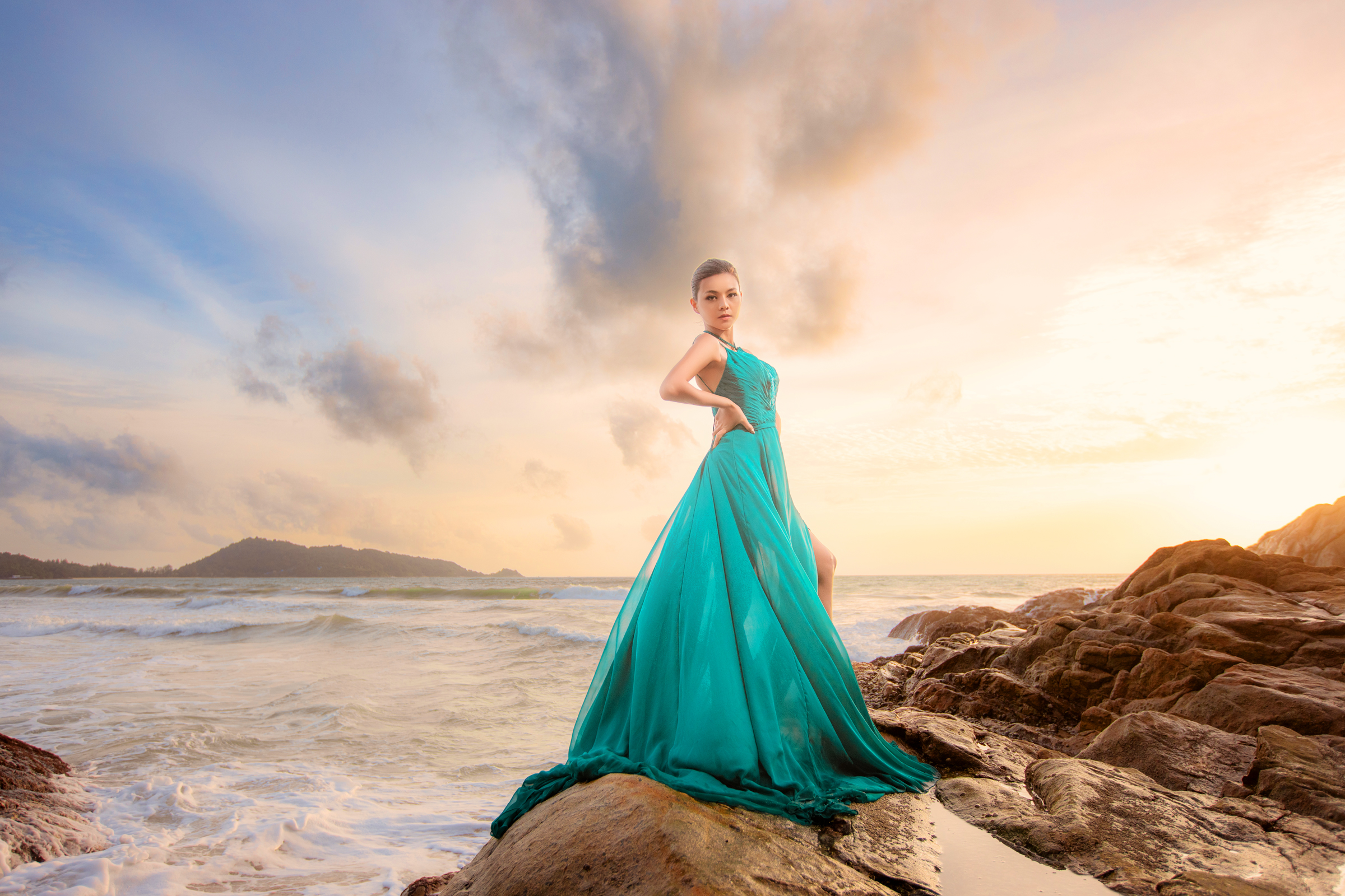 A model in a flowing turquoise dress poses confidently on a rocky beach during sunset, with waves crashing in the background.