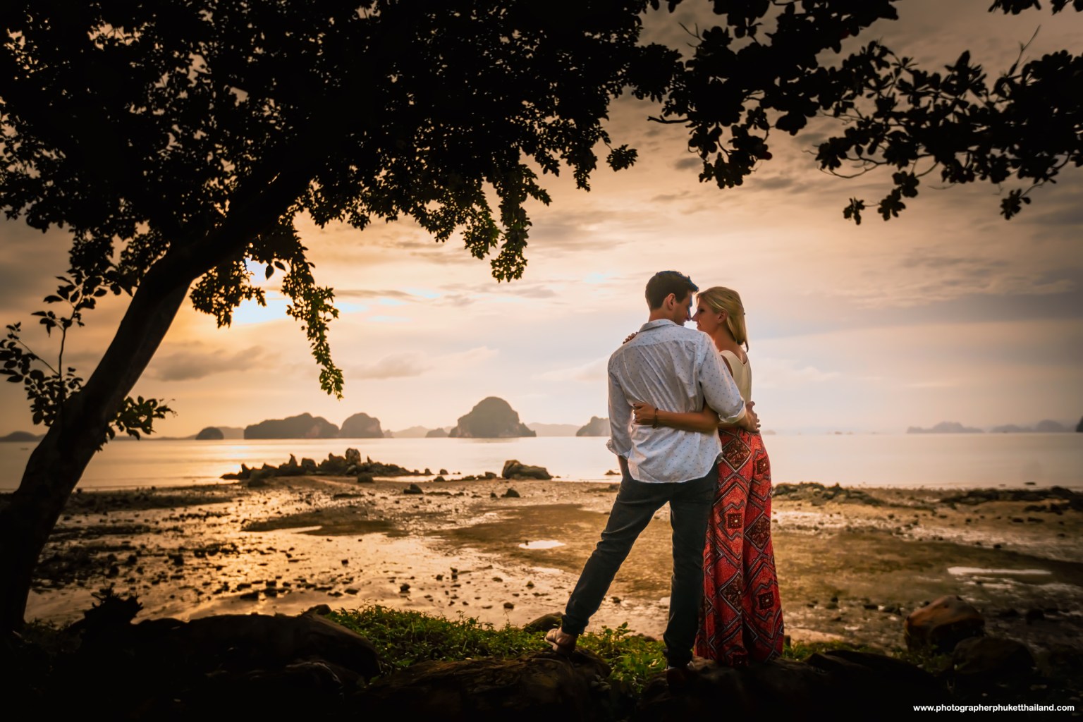 Couple embracing at sunset on a beach, surrounded by water and rocky islands in the background.