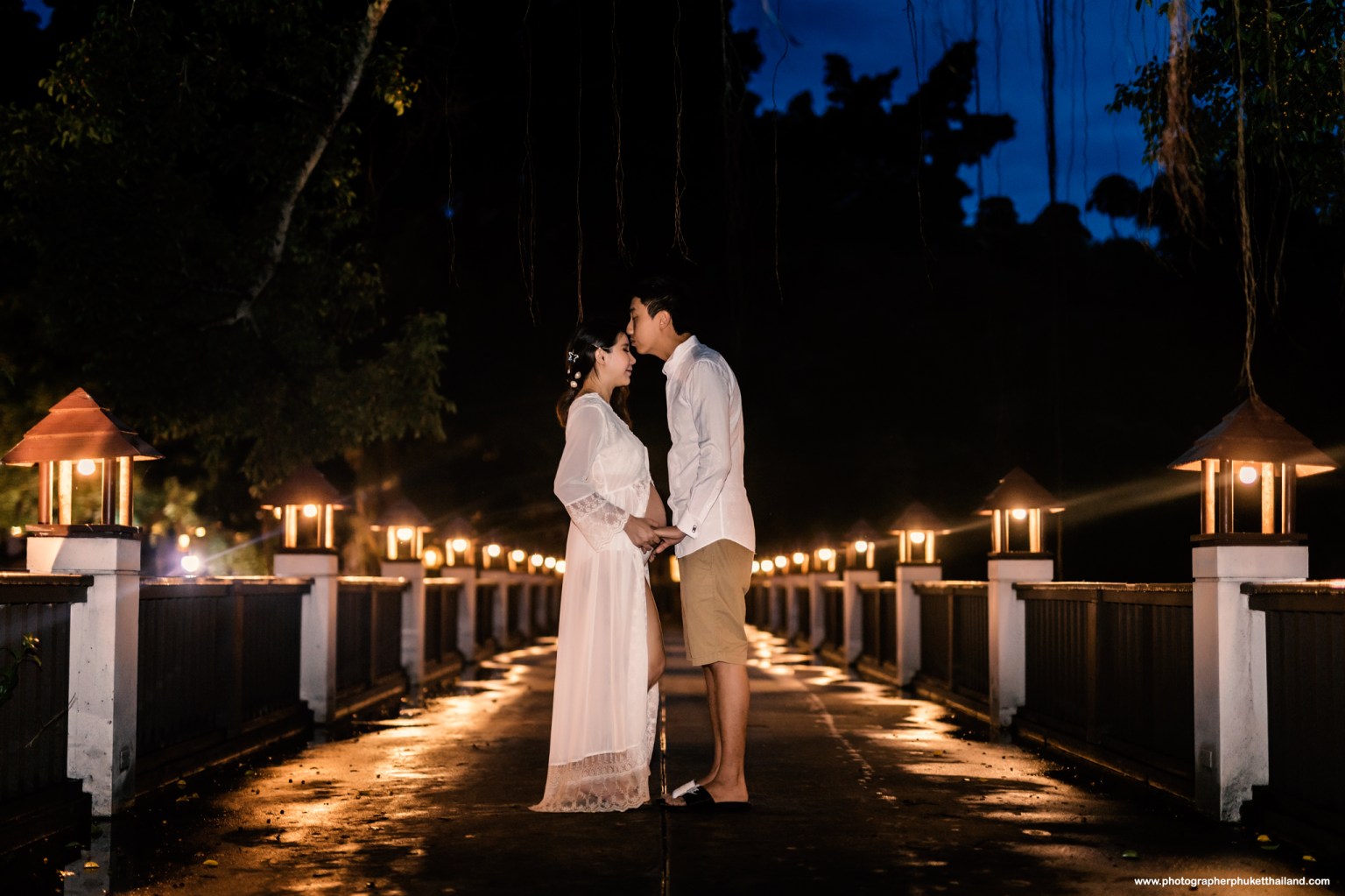 A couple stands on a beautifully lit walkway at night, expressing affection while holding hands. The scene features lanterns illuminating the pathway and a romantic atmosphere.