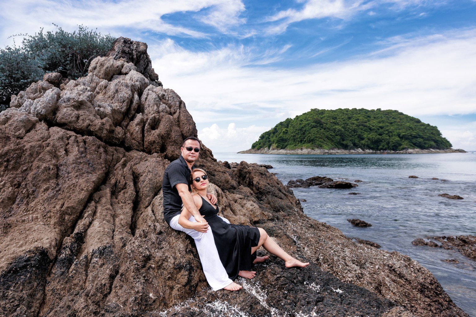 A couple sitting together on rocky shore, with a tropical island in the background and a blue sky with clouds.