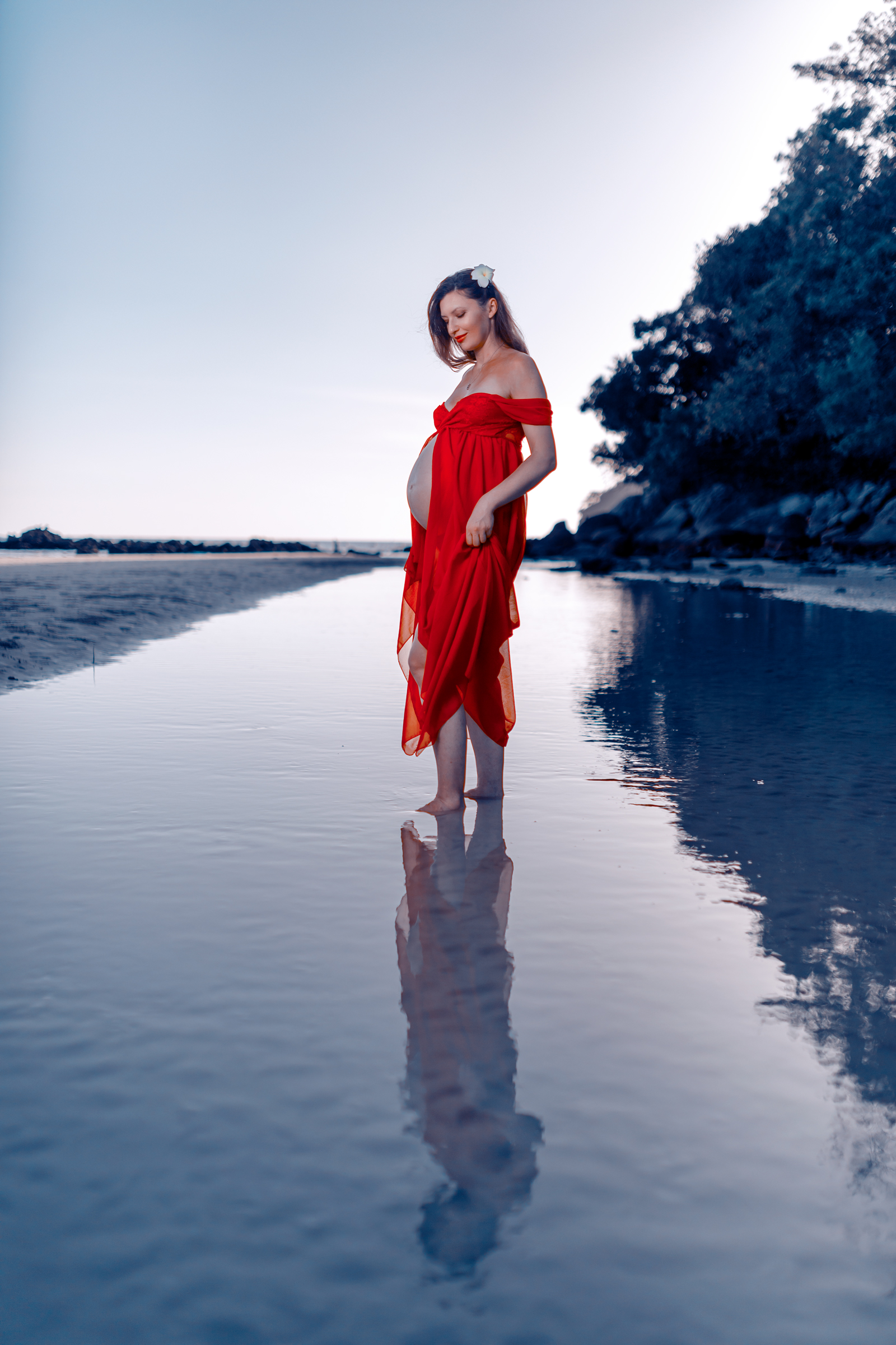 A pregnant woman in a flowing red dress stands barefoot in shallow water, reflecting the serene landscape and soft blue sky.