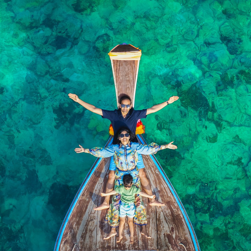 Aerial view of a family on a longtail boat in clear turquoise water, with two adults and a child joyfully posing with their arms outstretched.