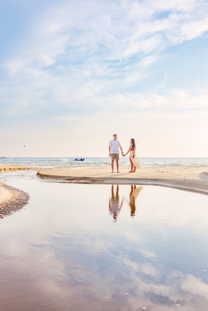 A couple holding hands on a beach at sunset, with reflections in the water and a serene sky.