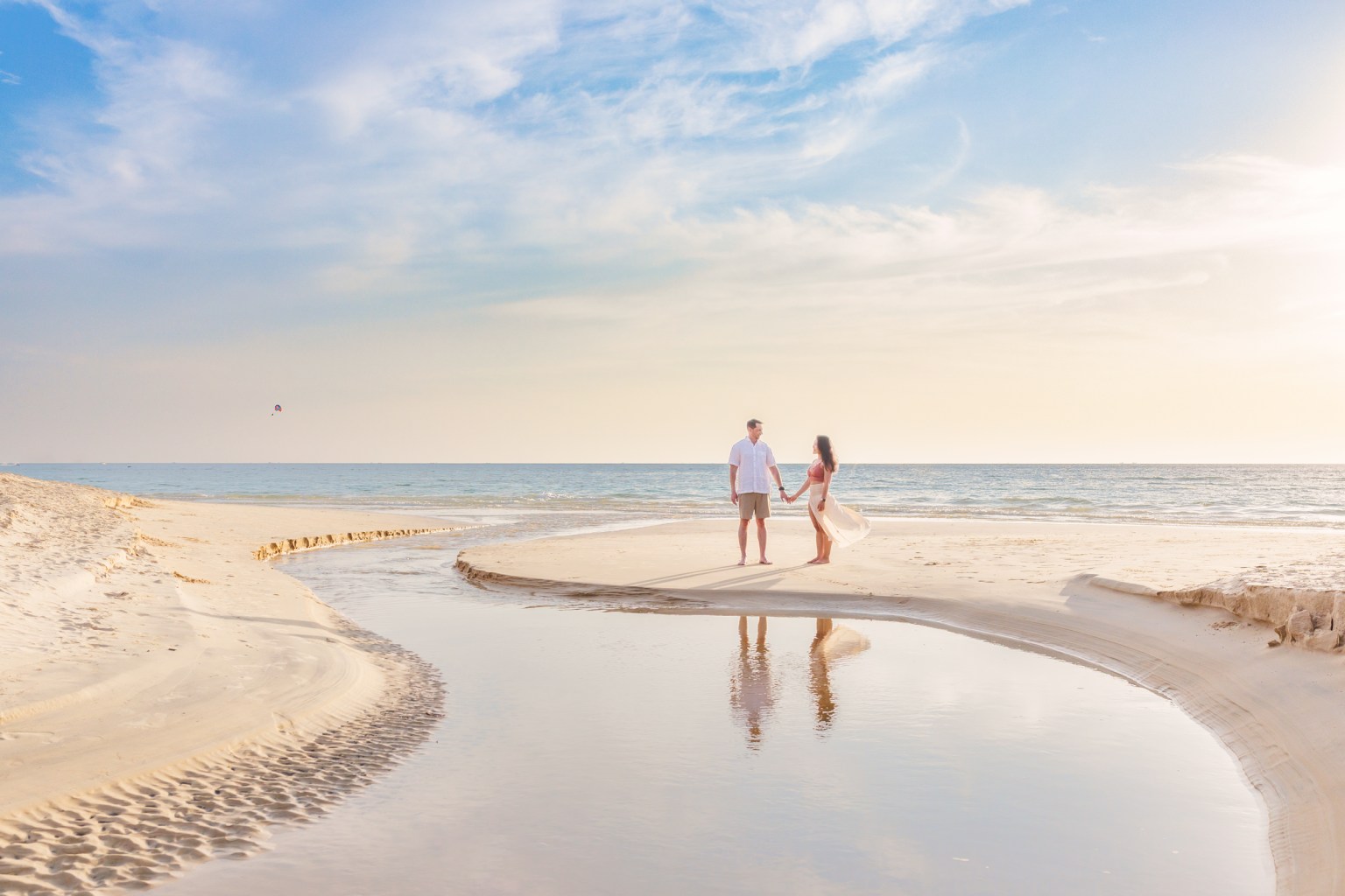 A couple stands hand in hand on a sandy beach during sunset, with a gentle stream of water reflecting their image.