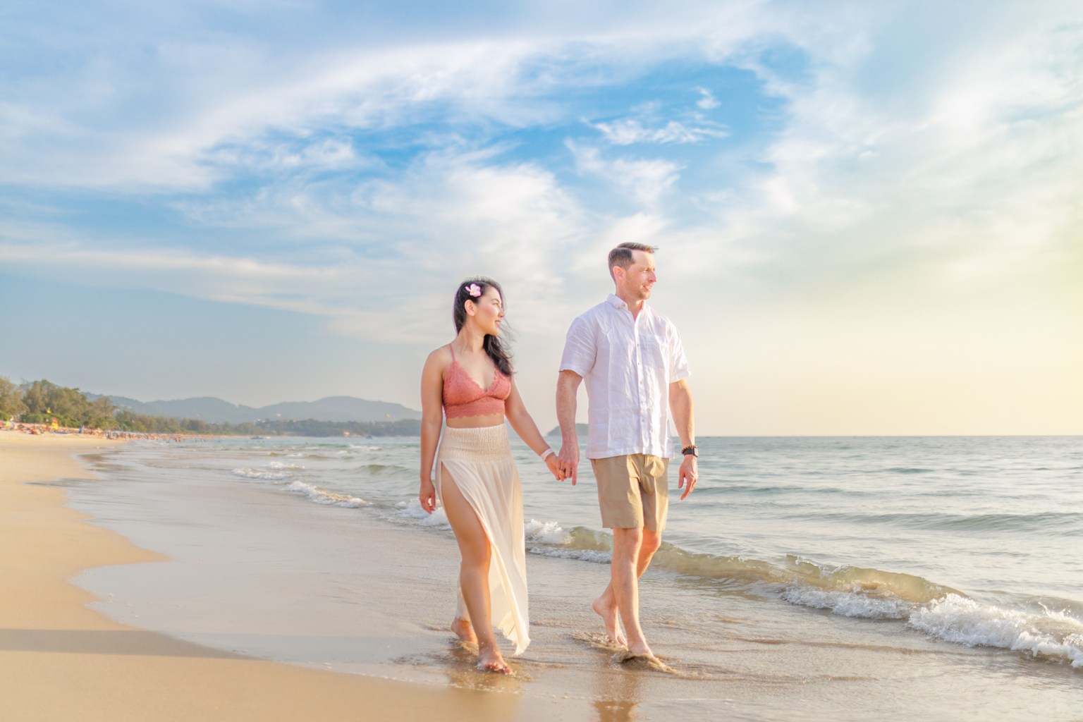 A couple walking hand in hand along the beach during sunset, with gentle waves lapping at their feet.