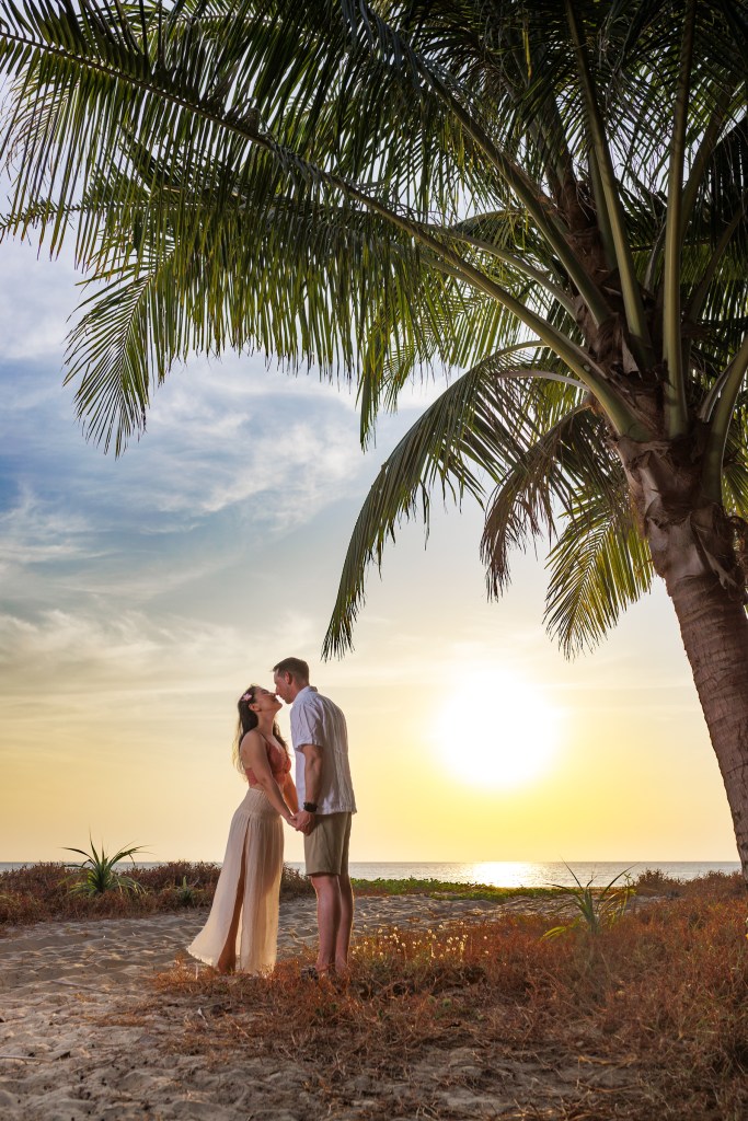 A couple embraces on a beach at sunset, framed by palm trees, capturing a romantic moment.