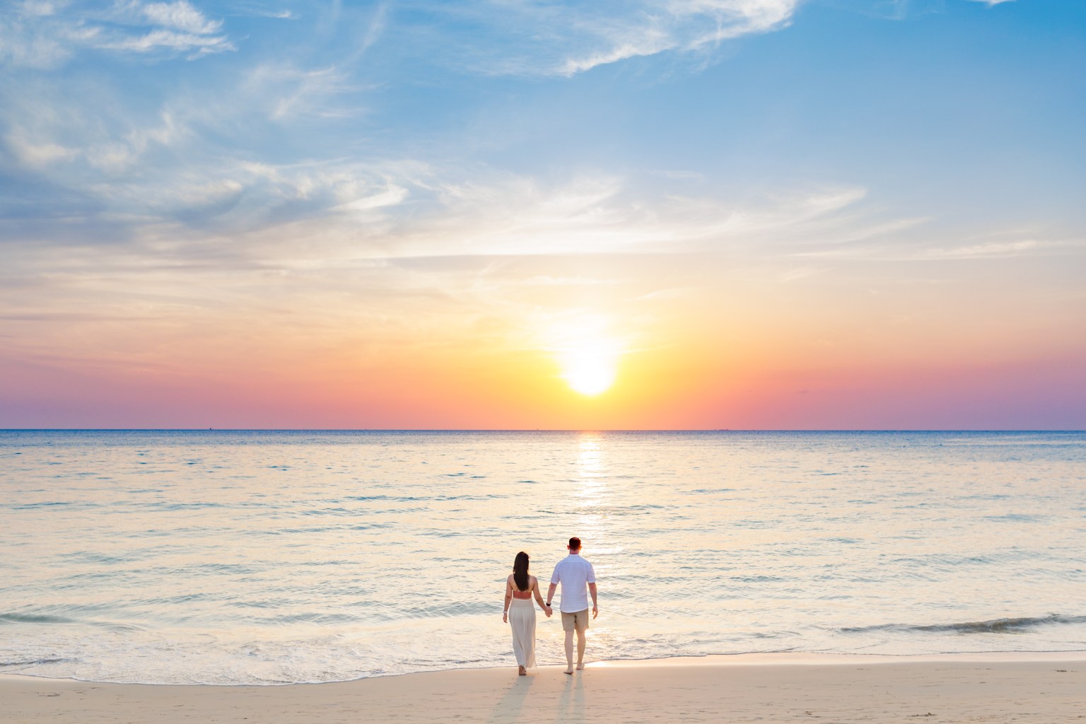 A couple walking hand in hand along a beach at sunset, with a vibrant sky displaying hues of orange, pink, and blue reflecting on the water.