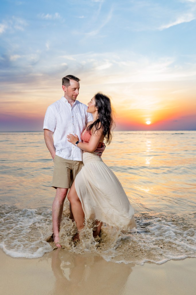 A couple enjoying a romantic moment on karon beach, standing in the water during sunset, with a colorful sky in the background.