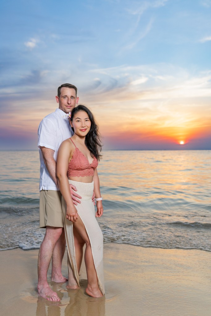 A couple standing on karon beach at sunset, with the ocean in the background, posing lovingly together.