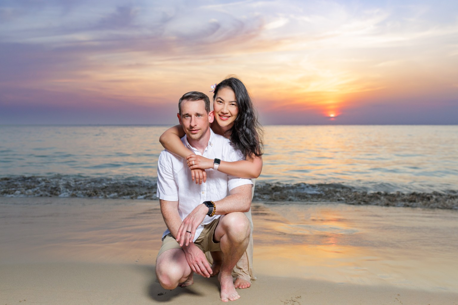 A couple poses on karon beach during sunset, with one partner sitting and the other embracing from behind.