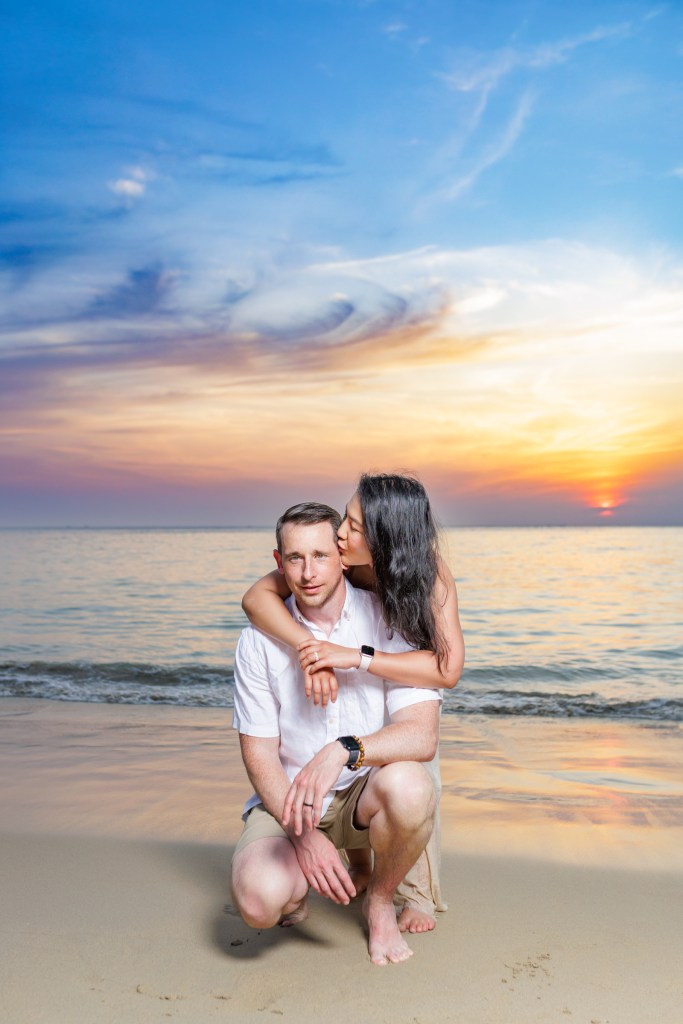 A couple embraces while sitting on the karon beach during sunset, showcasing a romantic moment against a colorful sky.