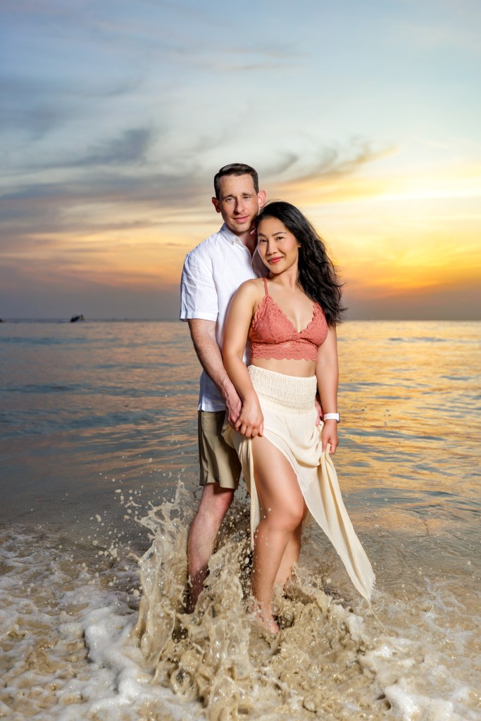 A couple standing in the water as sunset at karon beach phuket smiling and holding hands. The woman wears a pink top and light skirt, while the man is in a white shirt and beige shorts.