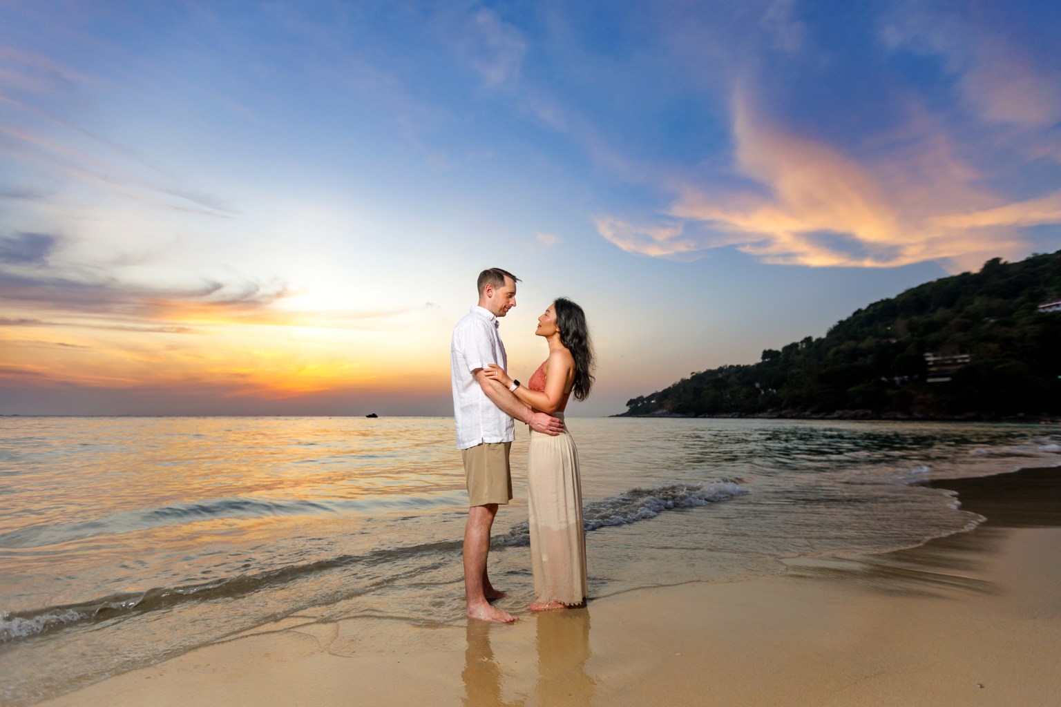 A couple standing on the beach, embracing each other at sunset. The sky displays warm hues of orange and purple, reflecting off the water at karon beach phuket