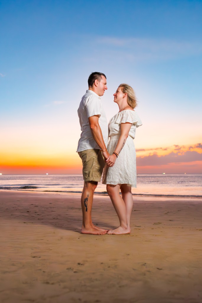 A couple holding hands on a beach during sunset, with a colorful sky and gentle waves in the background at karon beach phuket