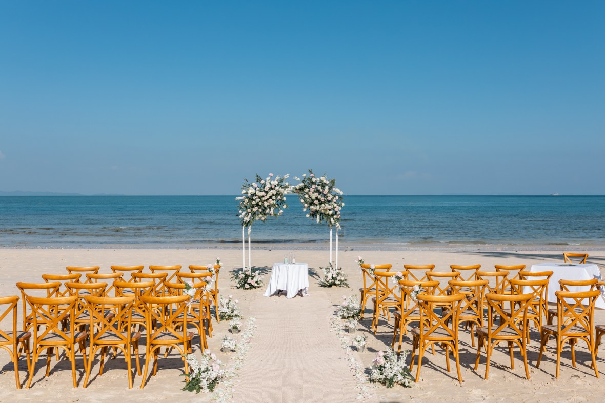 A beach wedding setup featuring rows of wooden chairs facing a floral arch and a small table. The backdrop includes a clear blue sky and calm ocean waves at cape panwa phuket