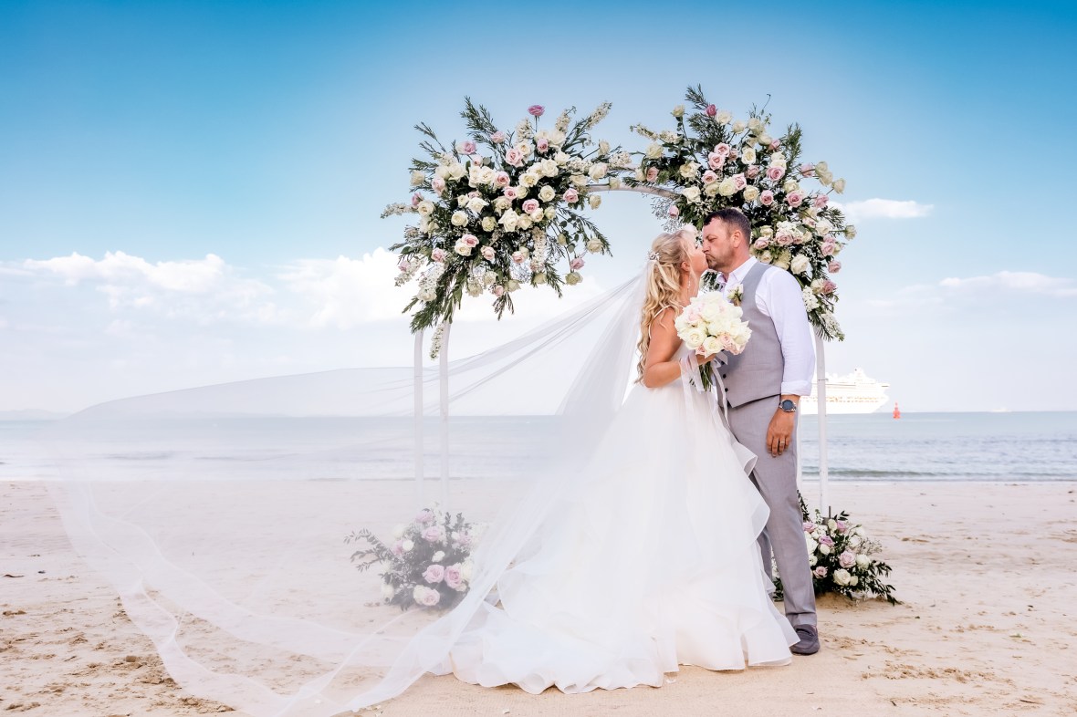 A bride and groom embracing under a floral arch on a beach during their wedding ceremony.