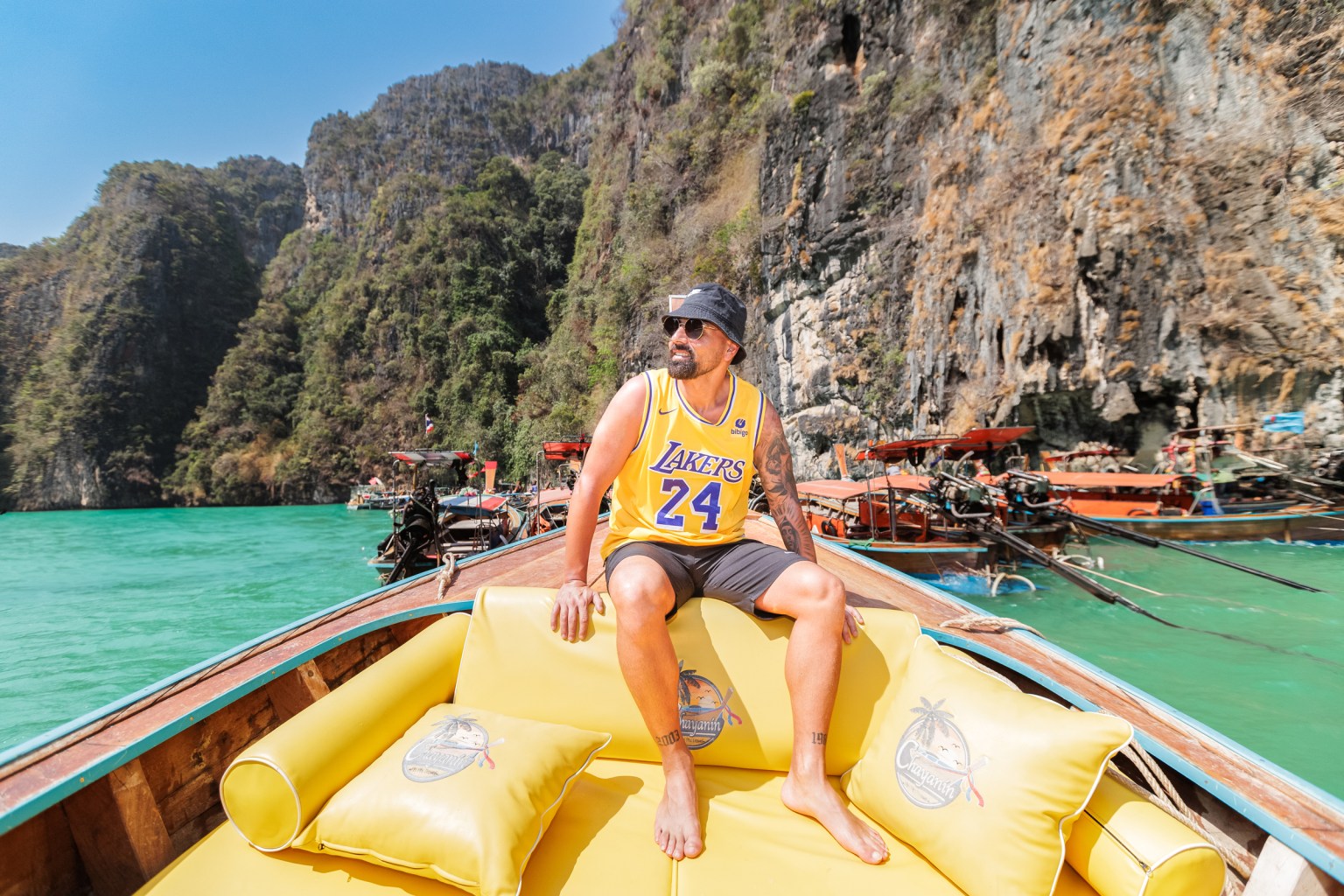 Man sitting on a yellow boat cushion, wearing a Lakers jersey and sunglasses, with rocky cliffs and boats in the background.