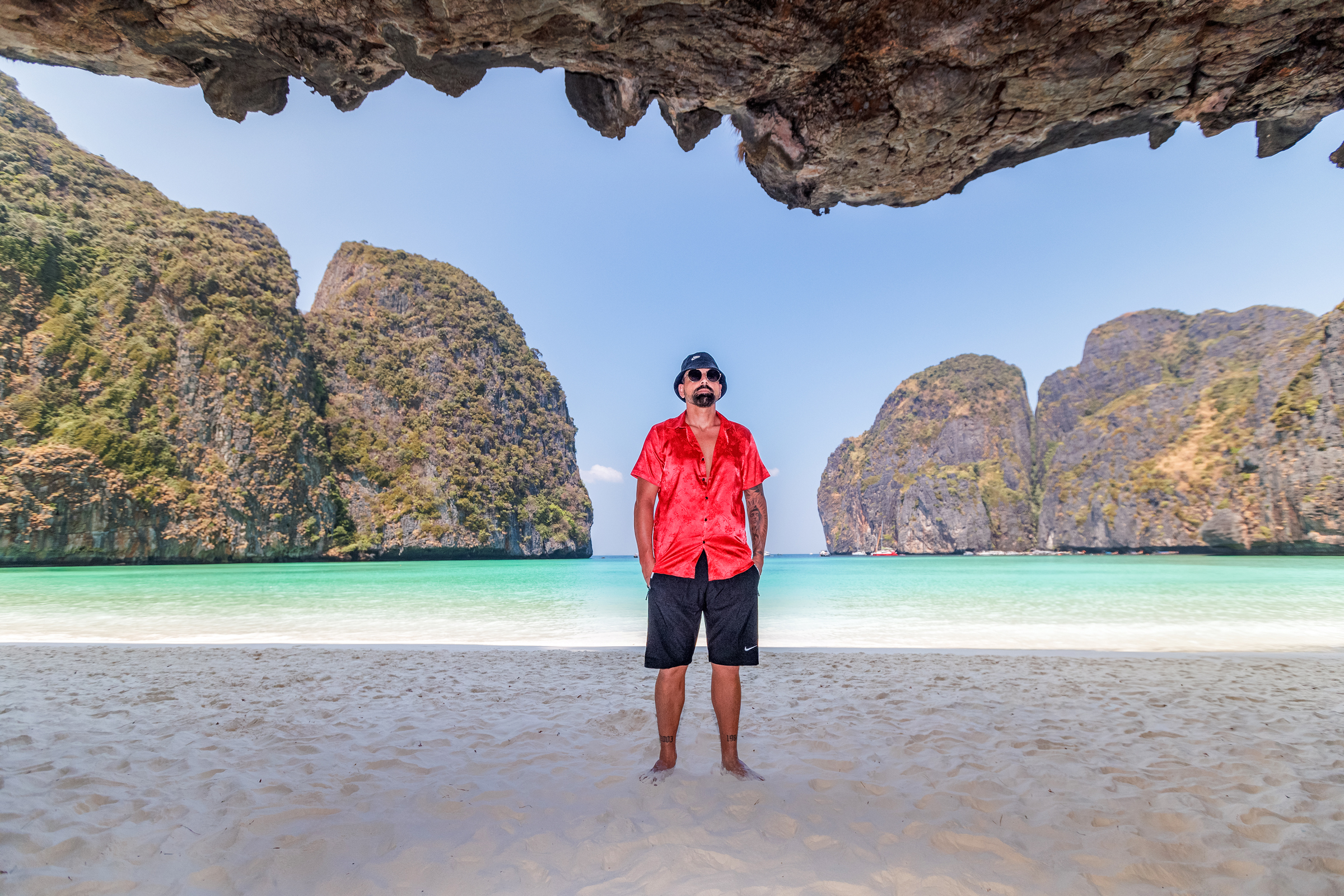 A man wearing a red shirt and sunglasses stands on a sandy beach, with green hills and clear turquoise water in the background.