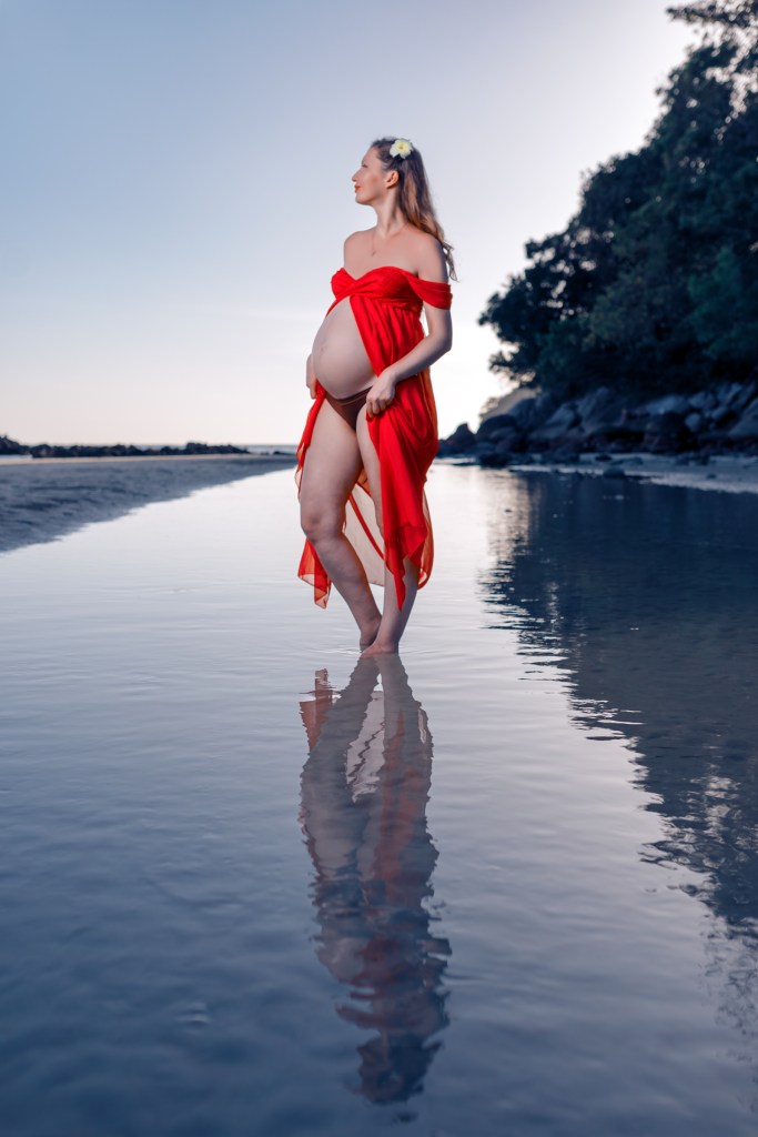 Pregnant woman in a flowing red dress standing on a beach with water reflecting her image, captured during sunset.