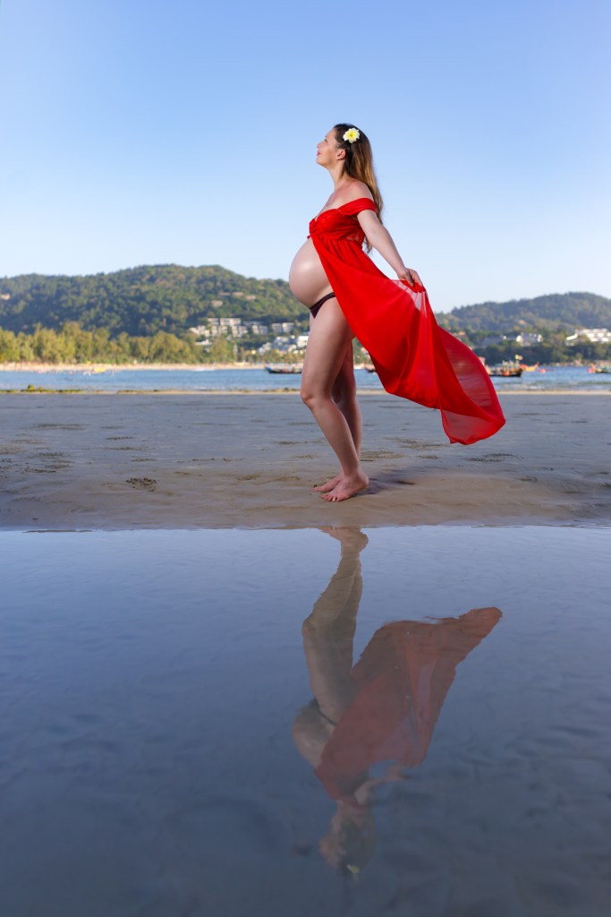 Pregnant woman in a red dress standing barefoot by the beach, with a beautiful landscape in the background and her reflection visible in the water.