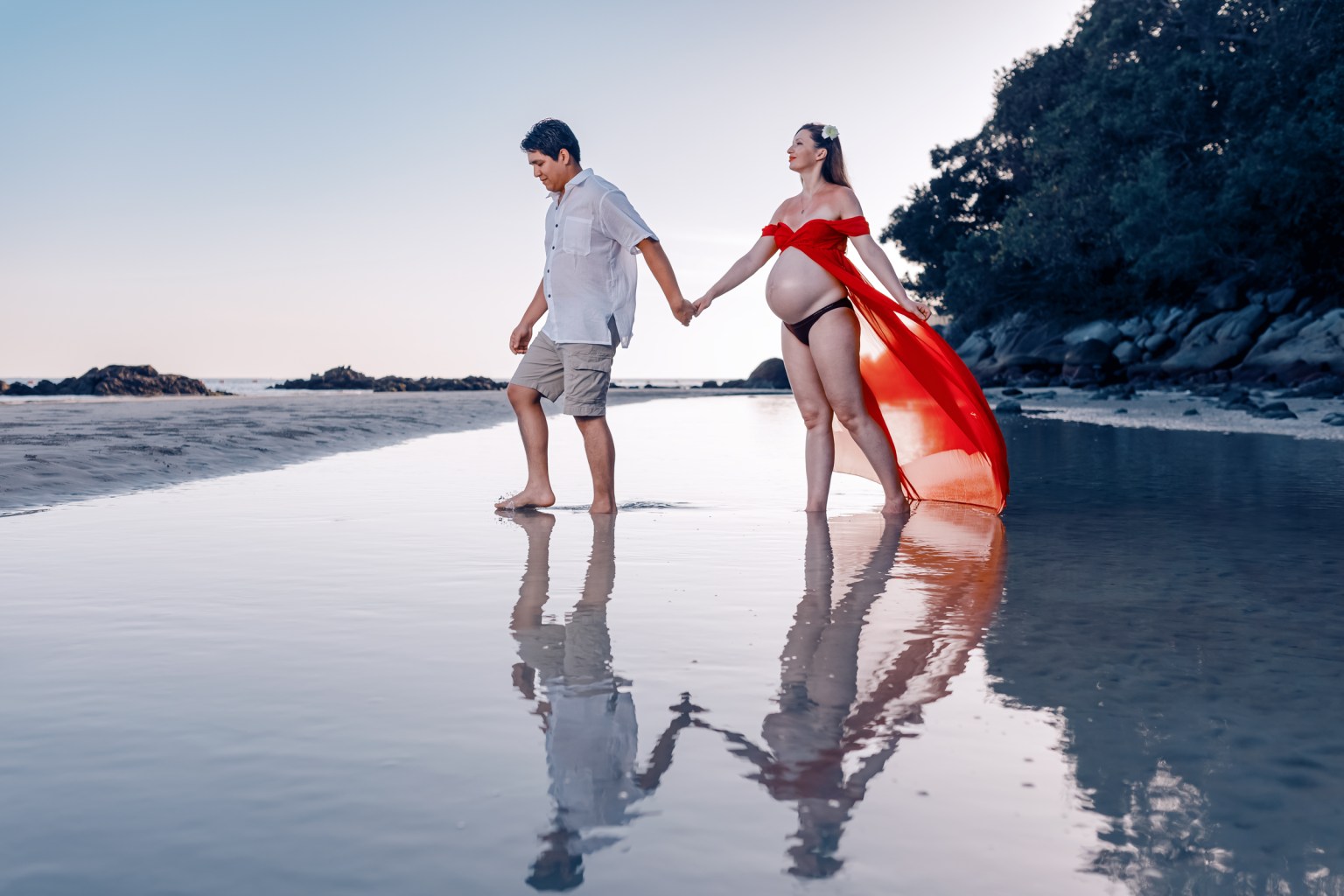 A pregnant woman in a flowing red dress and a man walking hand in hand along a beach at sunset, with their reflections visible in the wet sand.