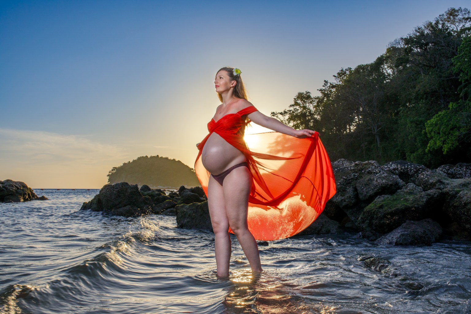 A pregnant woman in a flowing red dress stands in shallow water at the beach, with a bright sky and sun behind her.