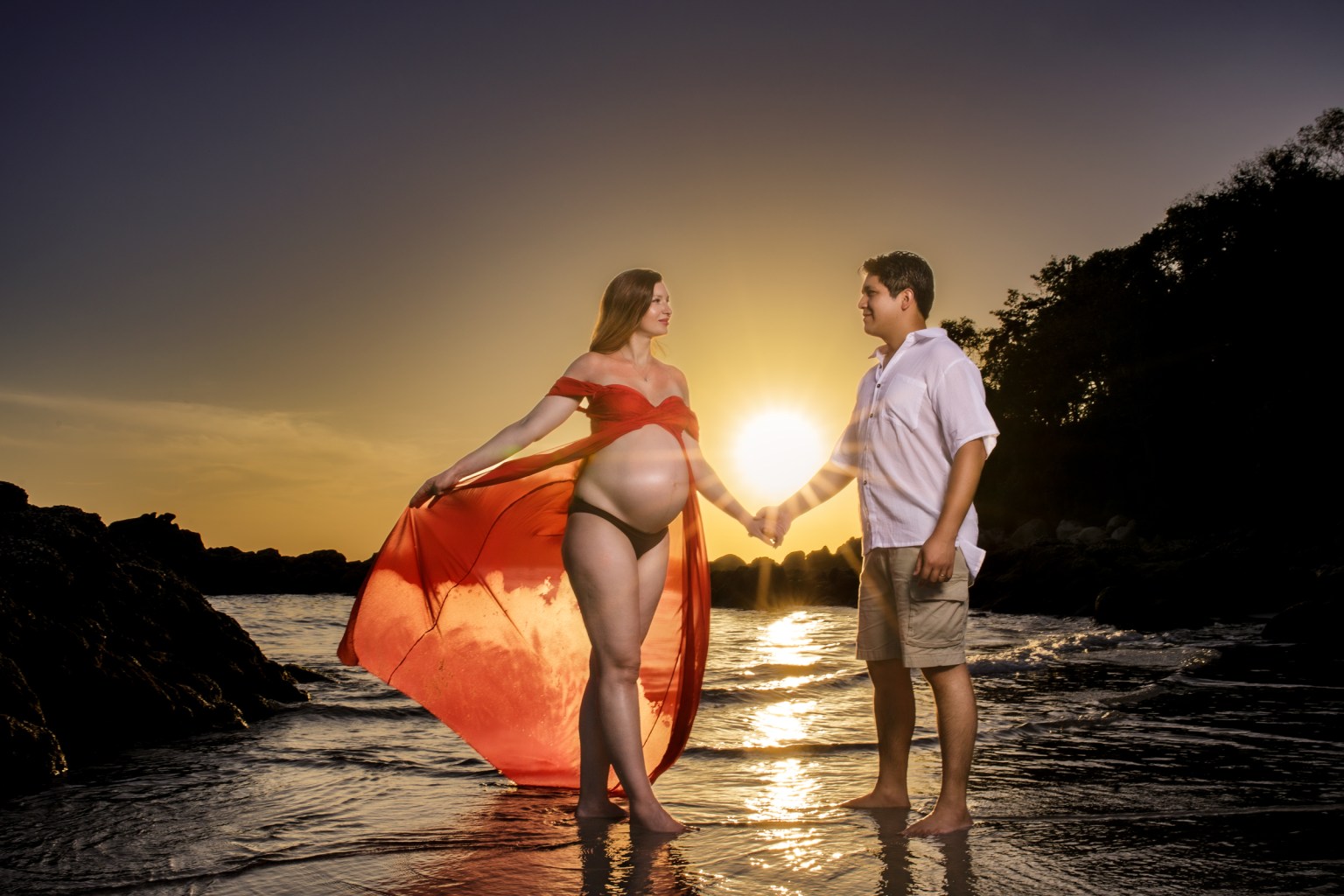A pregnant woman in a flowing red dress stands on a beach holding hands with a man, with the sun setting behind them.