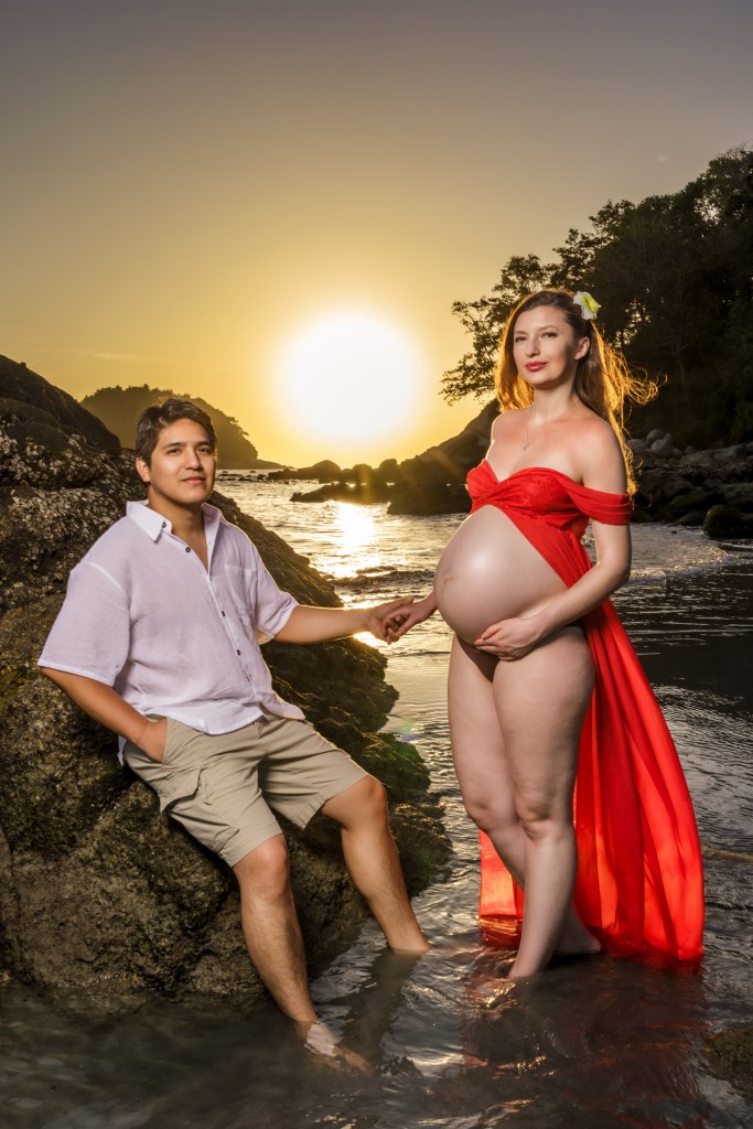 Couple posing on a rock by the beach at sunset, with the woman in a red dress accentuating her pregnancy.