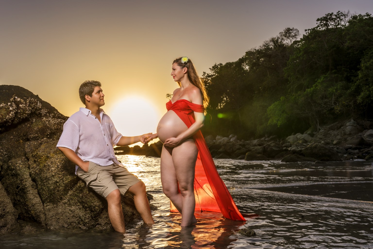 A pregnant woman in a red dress standing in the water holding hands with a man, with a sunset in the background.