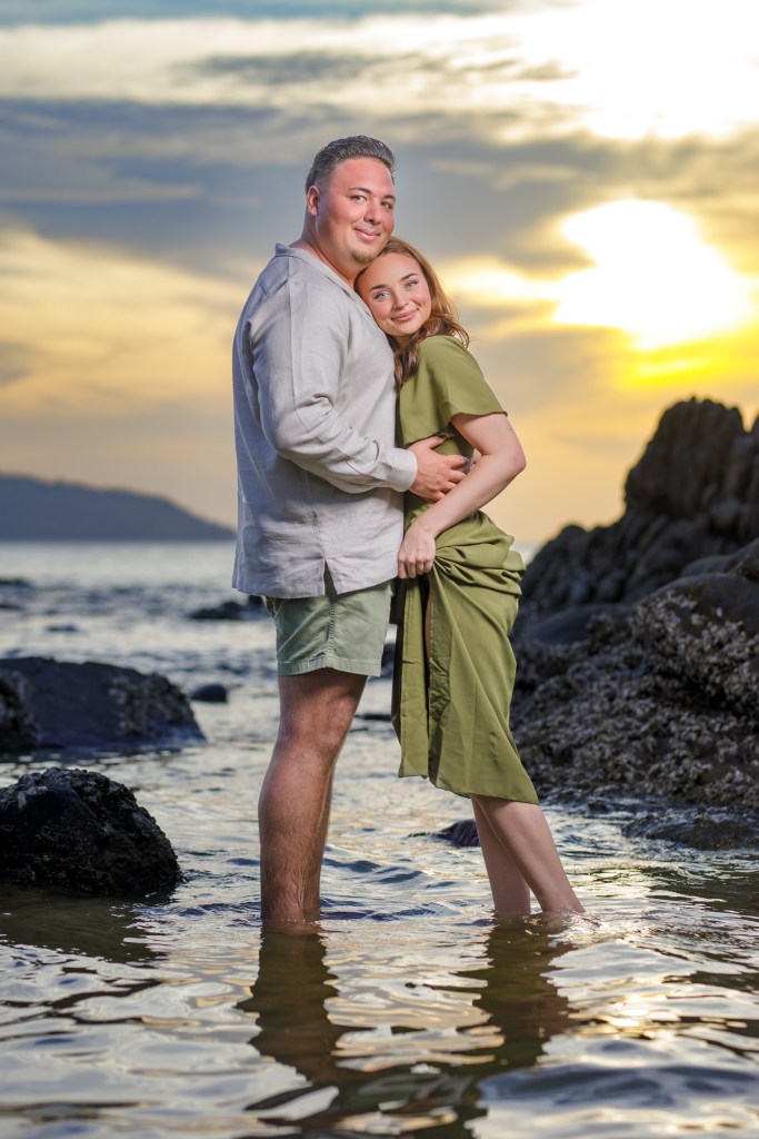 A happy couple embracing on a beach during sunset, standing in shallow water with rocky formations nearby.