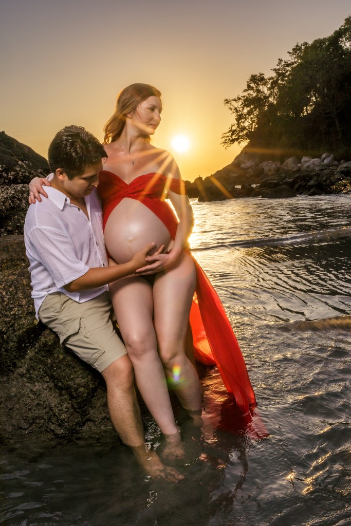 A couple enjoying a sunset photo shoot on the beach, with the pregnant woman wearing a flowing red dress and the man holding her belly.