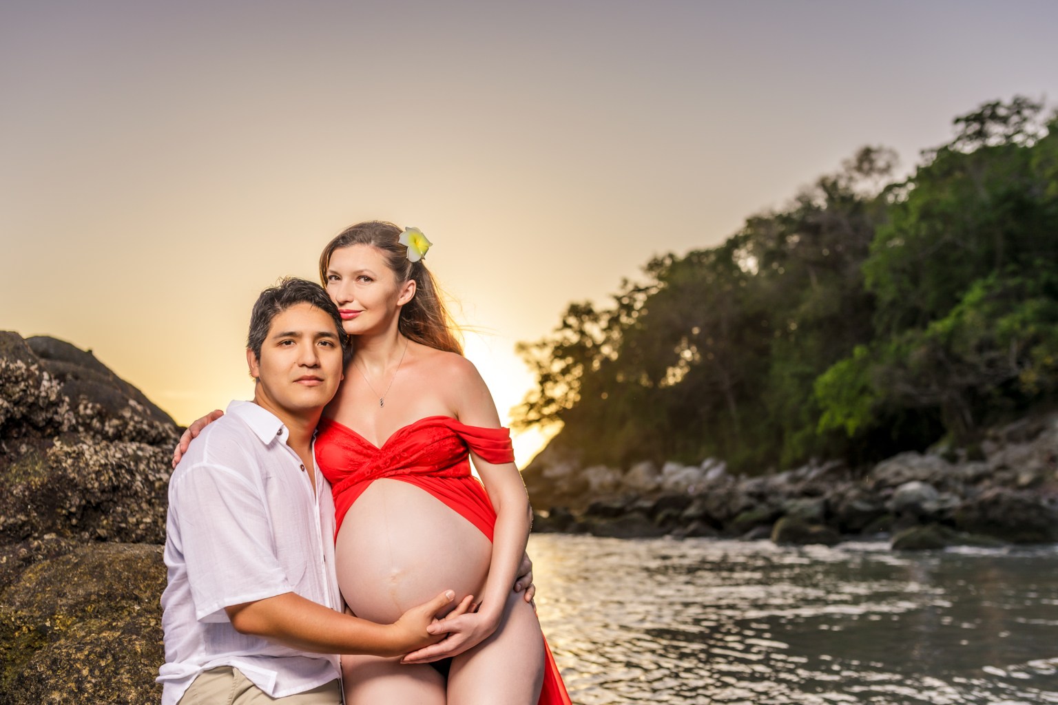 A pregnant woman in a red dress embraces her partner while sitting on rocks by the water at sunset, with a flower in her hair.