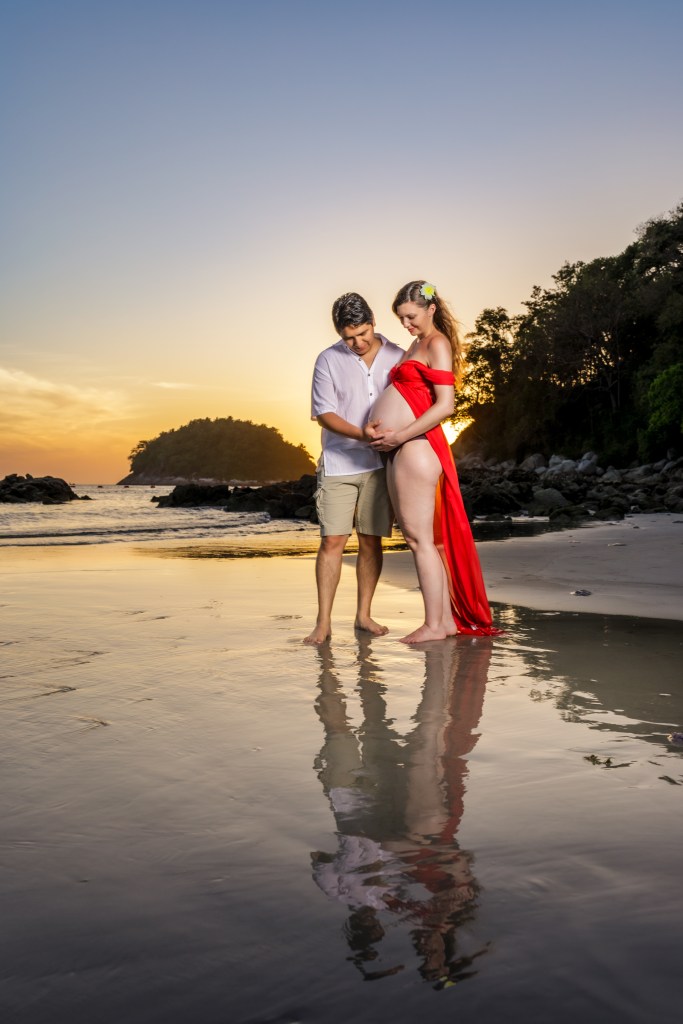 A couple stands on the beach during sunset, with the woman in a flowing red dress cradling her pregnant belly, while the man gently holds her waist. Their reflections can be seen in the wet sand.