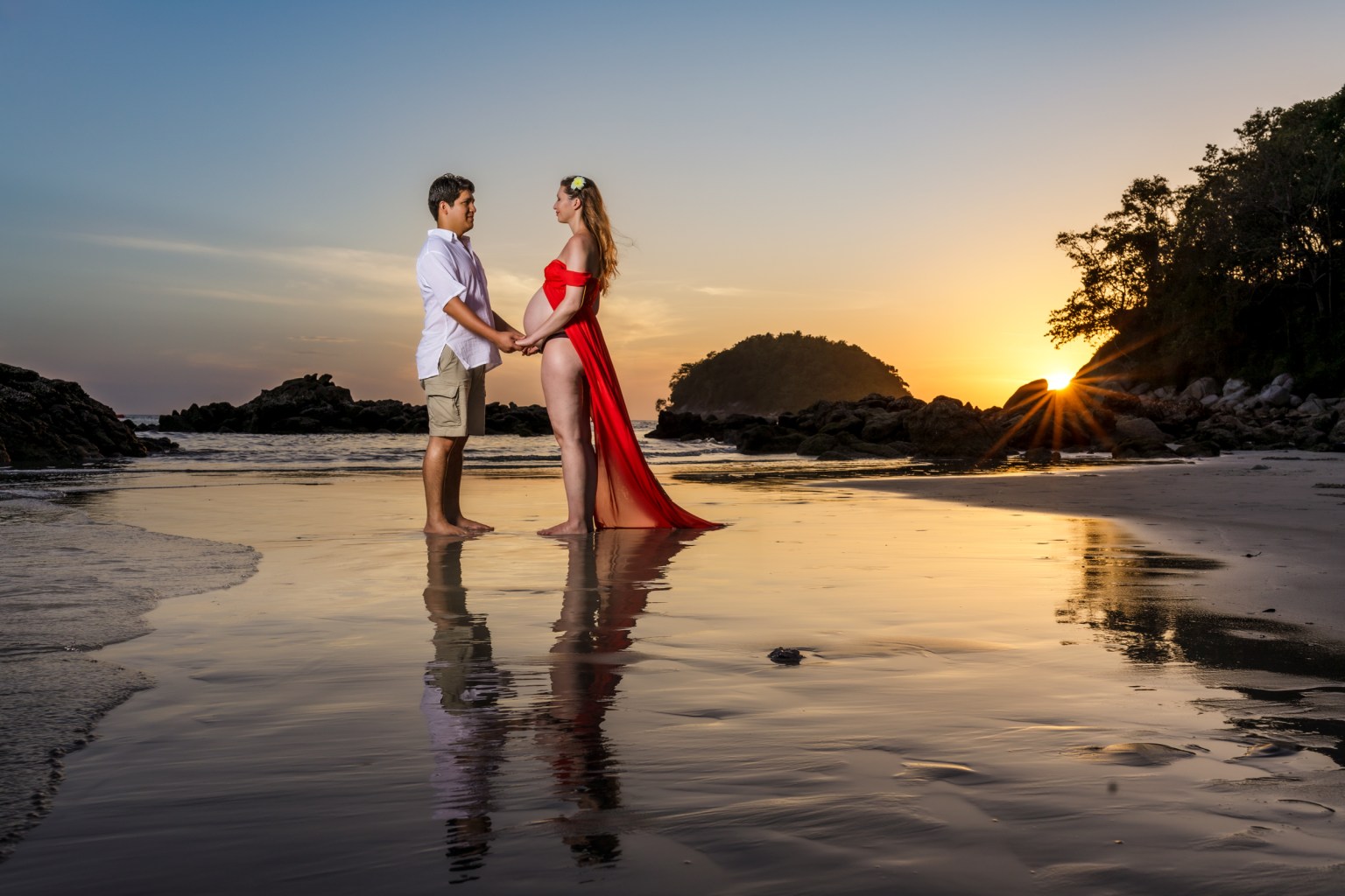 A couple standing on the beach during sunset, holding hands. The woman, pregnant, wears a flowing red dress, while the man is in a white shirt and shorts. Their reflections can be seen in the wet sand.