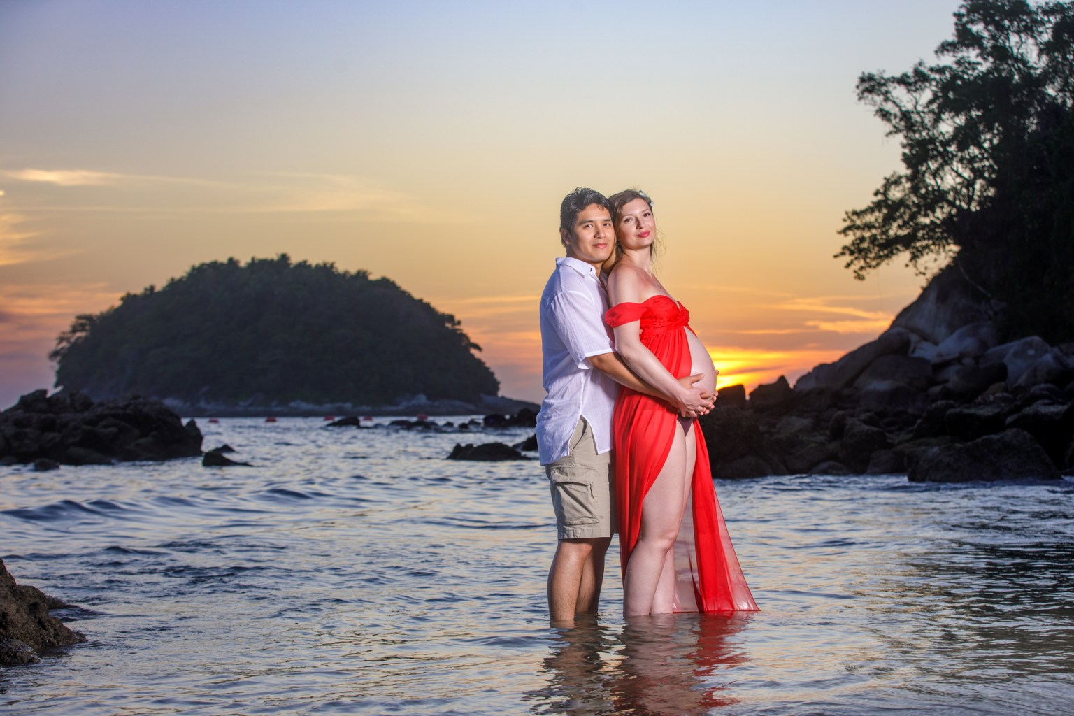 A couple standing in shallow water at the beach during sunset, with the woman wearing a flowing red dress and cradling her baby bump while the man stands beside her.