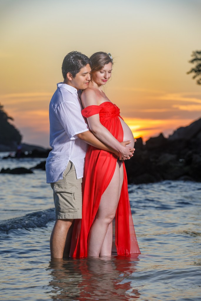 A couple standing together in the water at sunset, with the woman wearing a flowing red dress and the man in a white shirt and shorts.