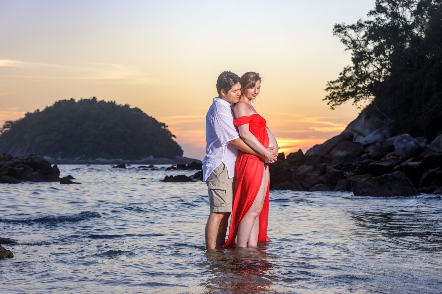 A couple standing in shallow water at the beach during sunset, the woman dressed in a flowing red gown and embracing her baby bump while the man gently holds her from behind.