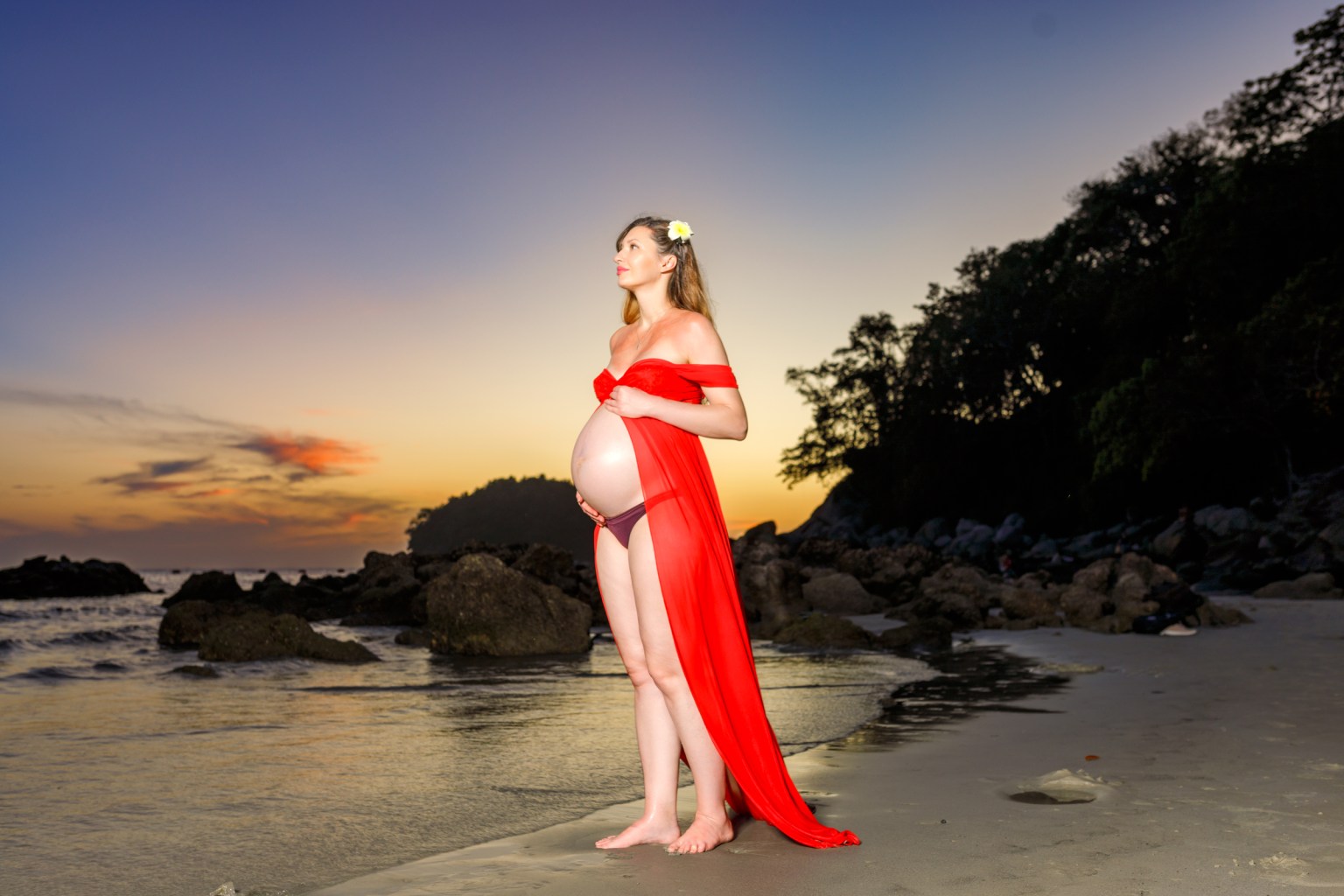 A pregnant woman in a red dress stands on a beach at sunset, gently holding her belly as she gazes at the horizon.