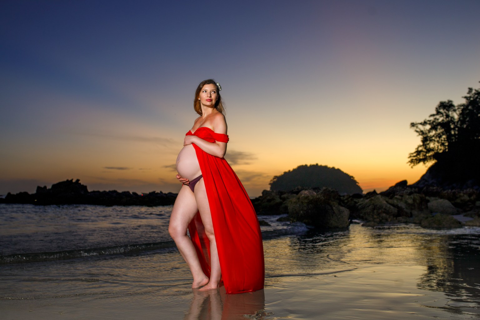 A pregnant woman wearing a red dress stands on a beach during sunset, with water reflecting her figure and a distant island silhouette in the background.