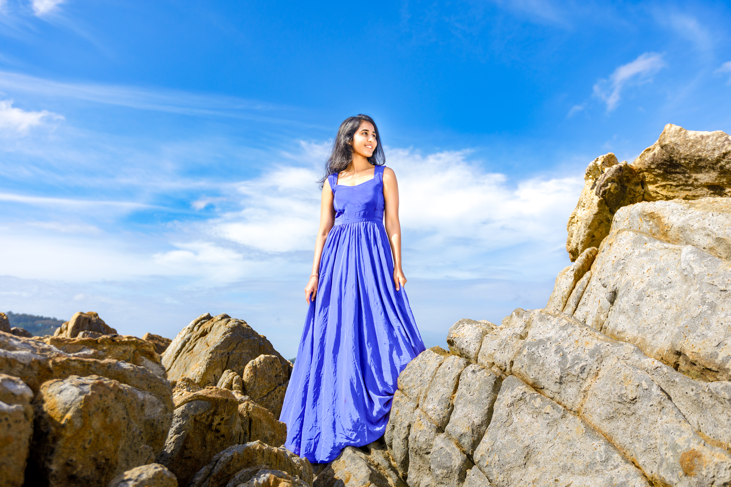 A woman wearing a blue dress stands on rocky terrain against a clear blue sky, showcasing a serene coastal landscape.