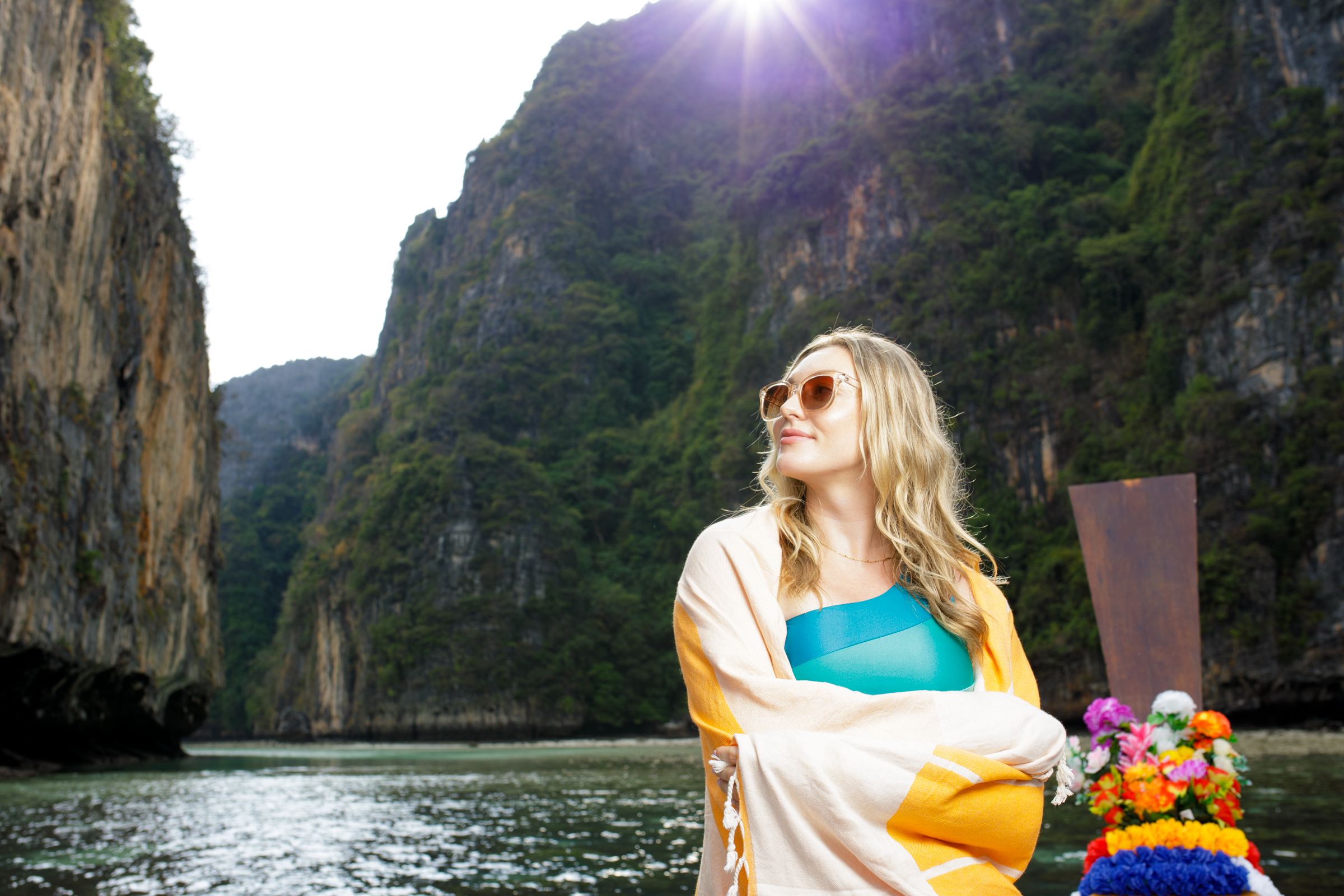 A woman wearing sunglasses and a colorful wrap stands on a boat, posing against a backdrop of towering cliffs and a bright sun in the background.