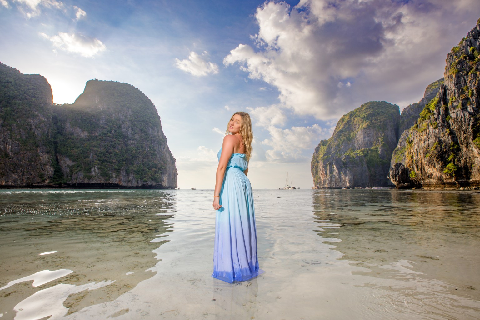 A model wearing a gradient blue dress poses in shallow waters at a scenic beach, surrounded by dramatic cliffs and a beautiful sky.