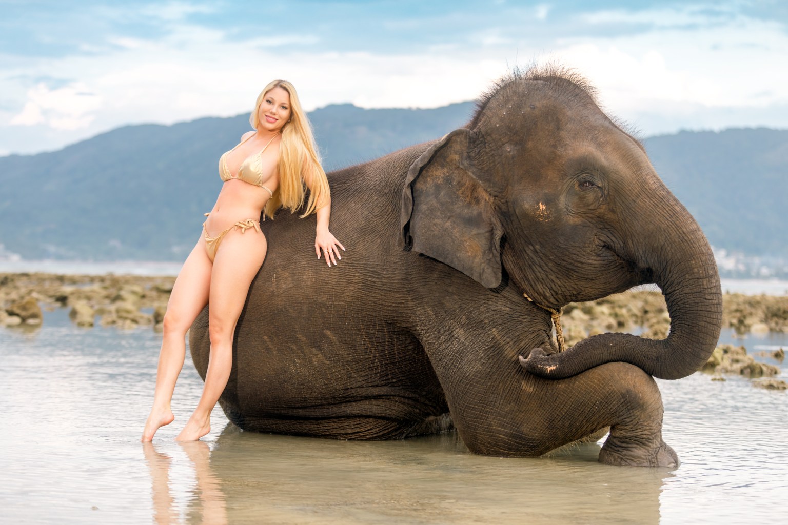 A woman in a bikini poses beside an elephant in shallow water, with a mountainous background.