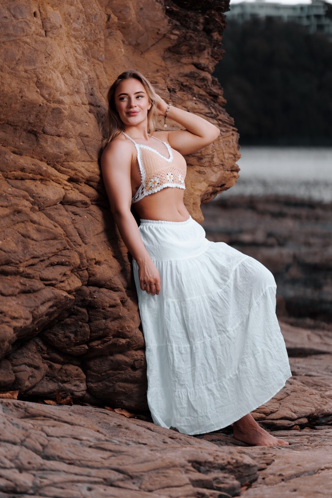 A young woman poses against a rocky backdrop, wearing a crochet top and a long white skirt. The scene captures a natural and relaxed vibe, highlighting her casual yet stylish look.
