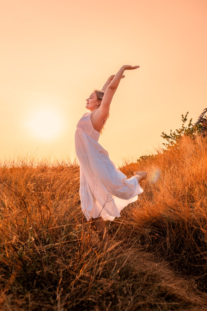 A woman in a white dress joyfully stretches her arms overhead while standing on a grassy hill at sunset, with warm orange light illuminating the scene.