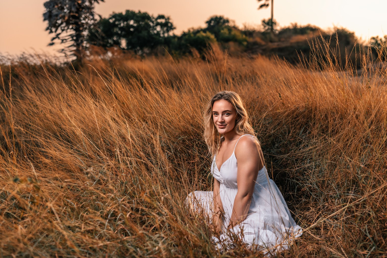 A woman sitting in tall golden grass during sunset, wearing a white dress and smiling.