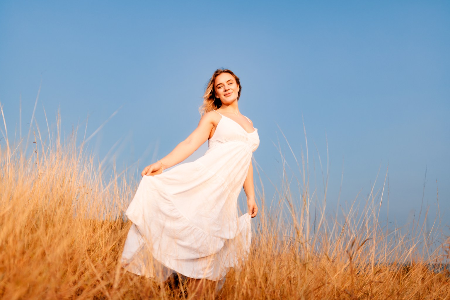 A woman in a flowing white dress stands in tall grass against a clear blue sky, smiling and gently swaying her dress.